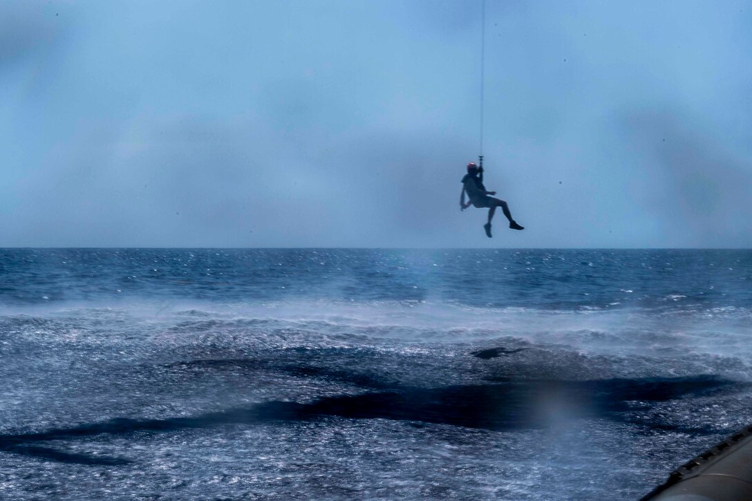 A service member hangs from a helicopter above water.