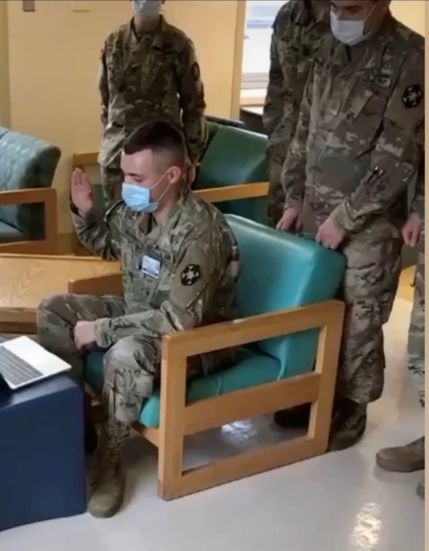 Soldier wearing a face mask and faces a laptop computer with his right hand raised as other soldiers stand behind him.