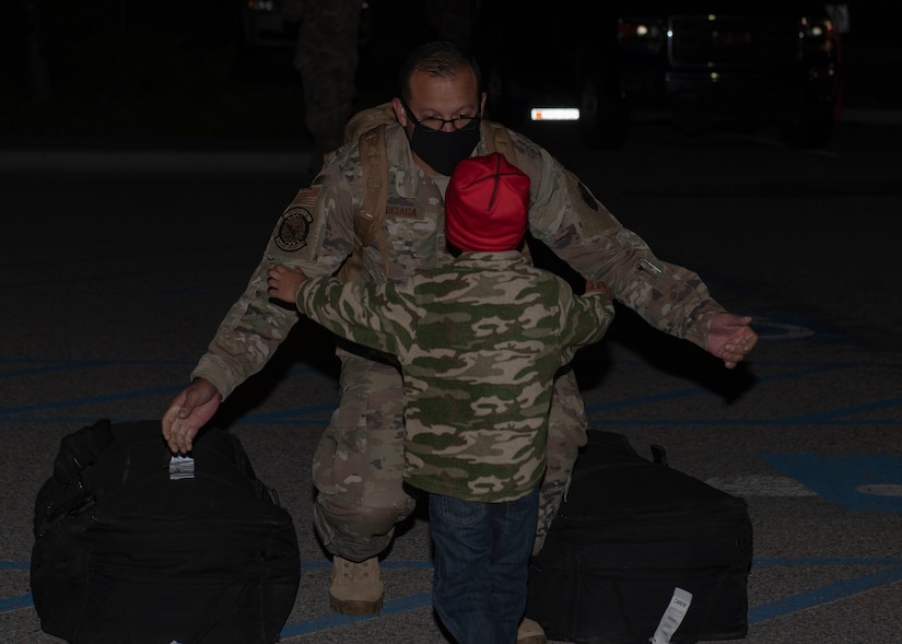 An airman wearing a mask and standing between two duffel bags crouches and opens his arms to hug his approaching son.