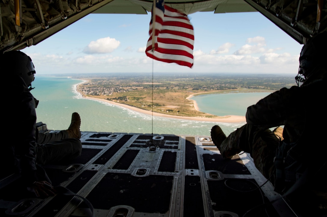 Two airmen, shown from behind in silhouette, look out over coastline from an open aircraft.