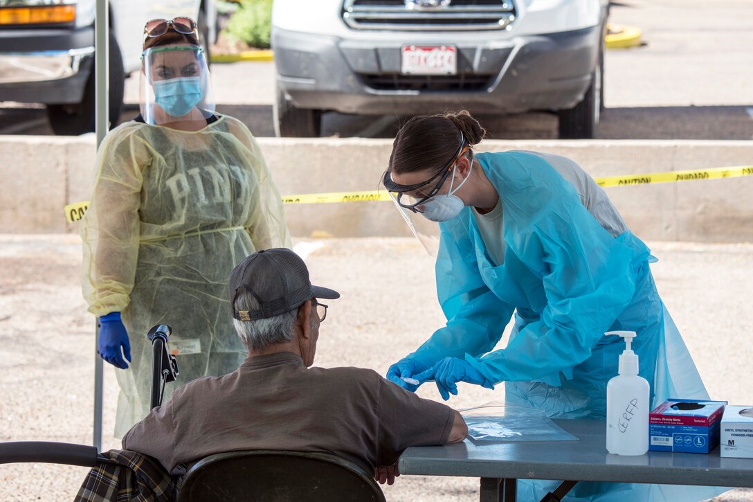 A service member in personal protective equipment prepares to administer a medical test to a nursing home resident. Another medical technician in PPE stands by.