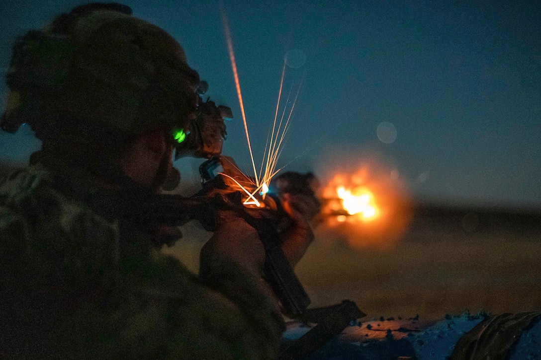 A soldier wearing a helmet and night vision optic fires a weapon at night.