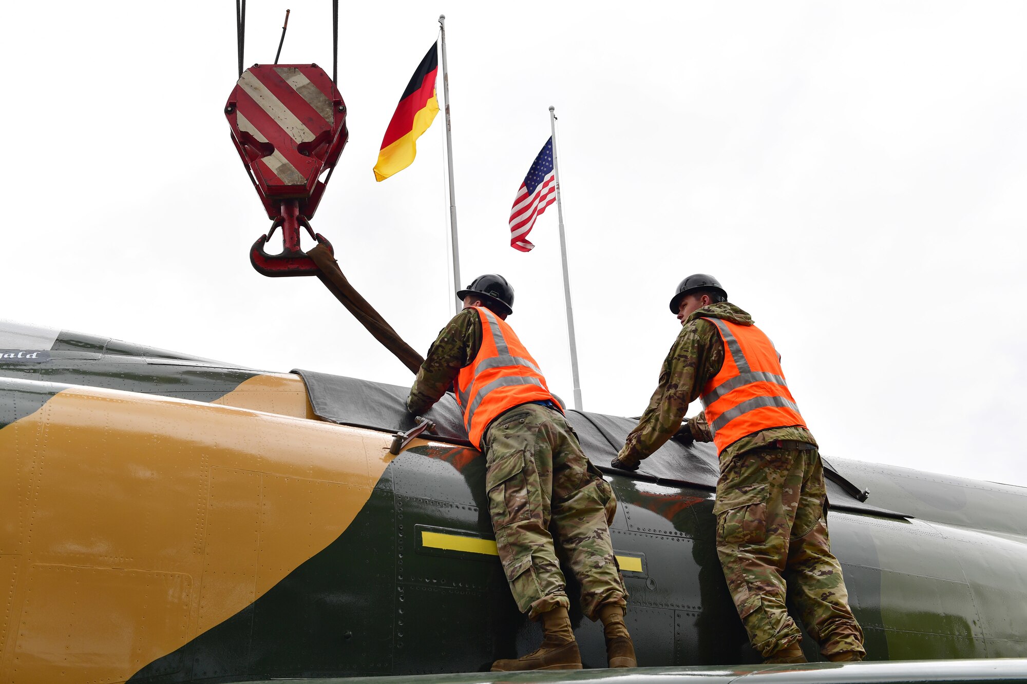 Two Airmen connect a crane load block to a jet aircraft.