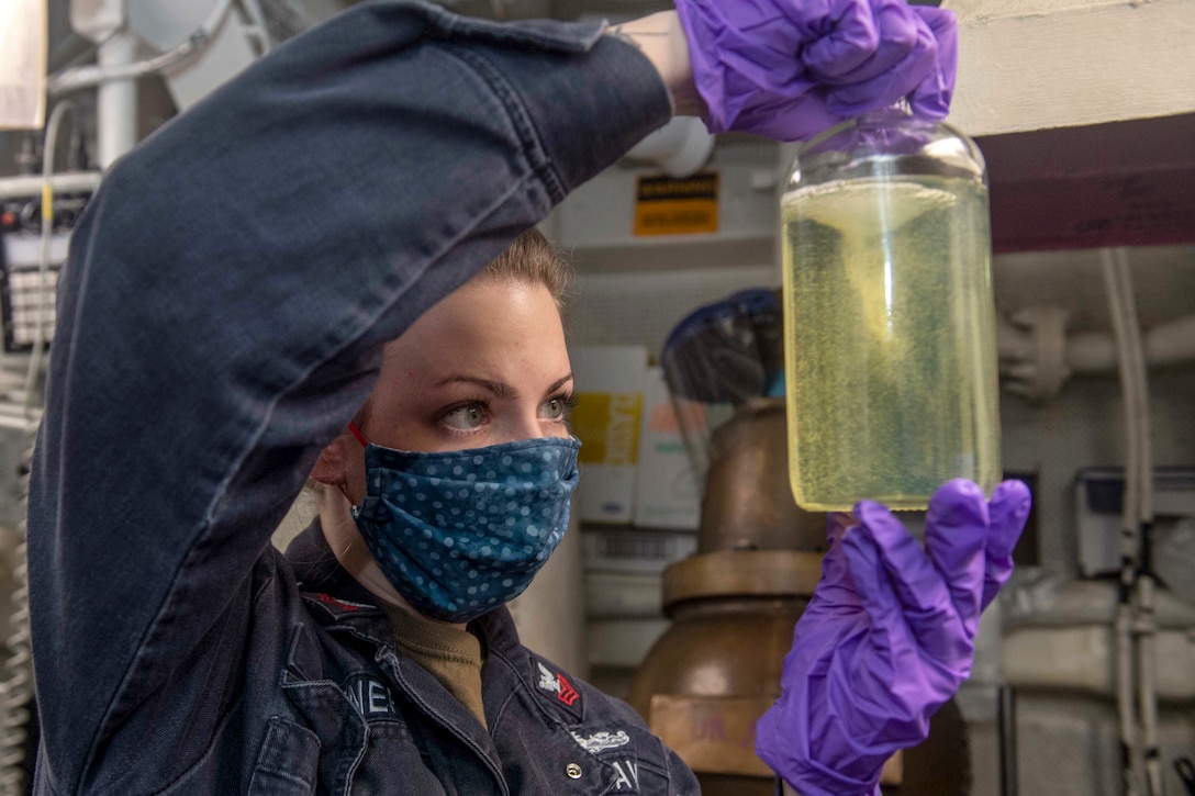 A sailor holds up a jar filled with yellowish liquid and looks at its contents.