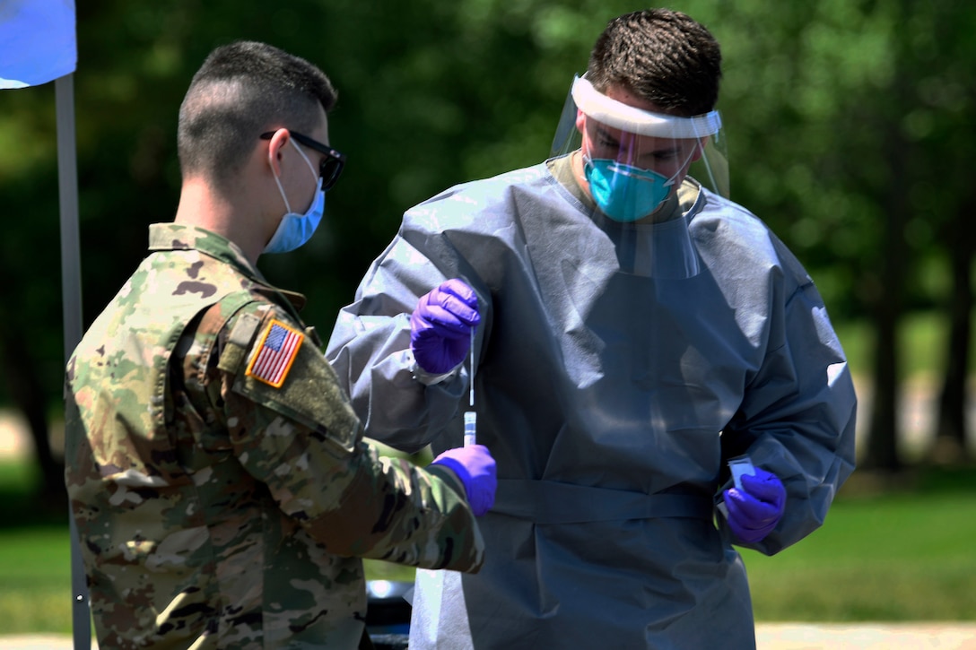 A National Guardsman wearing personal protective equipment puts a COVID-19 test swab in a test tube held by a person in a uniform who is wearing a mask and gloves.