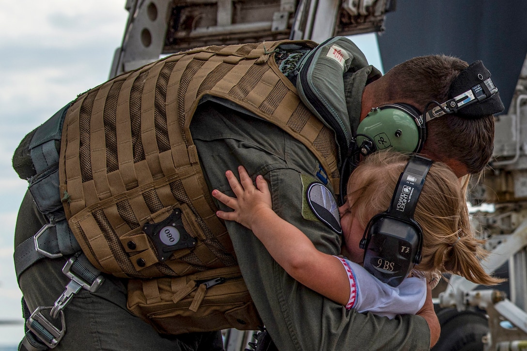 An airman wearing headphones hugs a young girl on a flightline, who is also wearing headphones.