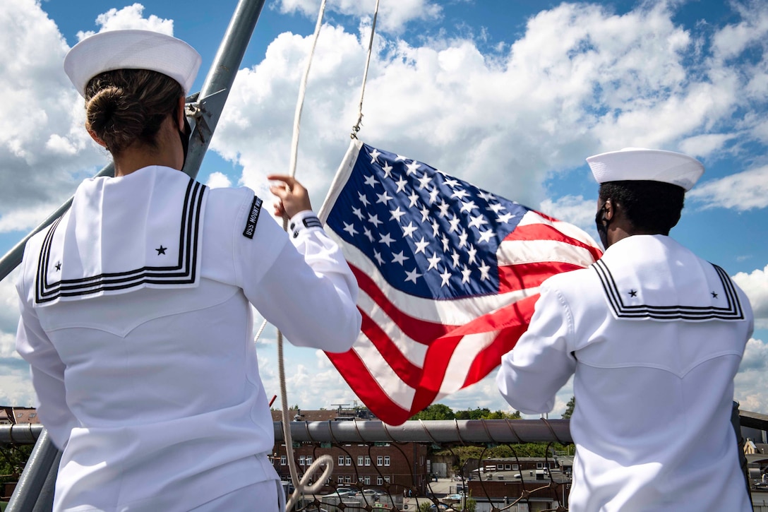 Two sailors raise an American flag on a ship’s mast.