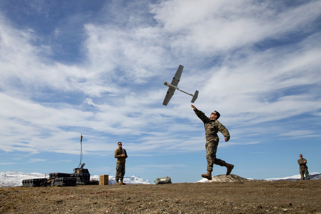 A Marine throws a winged drone into the sky as other troops watch in the background on flat, dirt terrain.