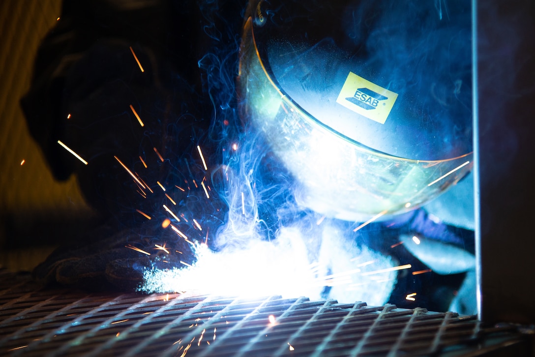 Blue smoke and sparks fly as an airman welds two parts together.