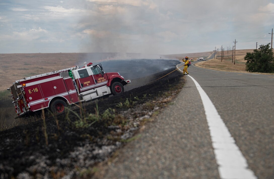 With smoke in the background, an airman directs an emergency vehicle up a slope onto the road.