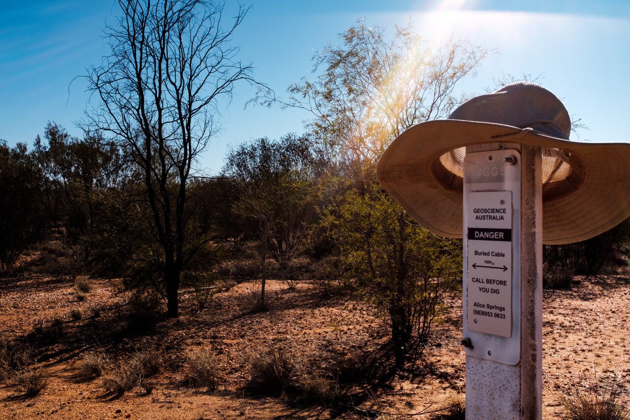 A wide-brimmed hat hangs on a warning sign in a desert area with trees.