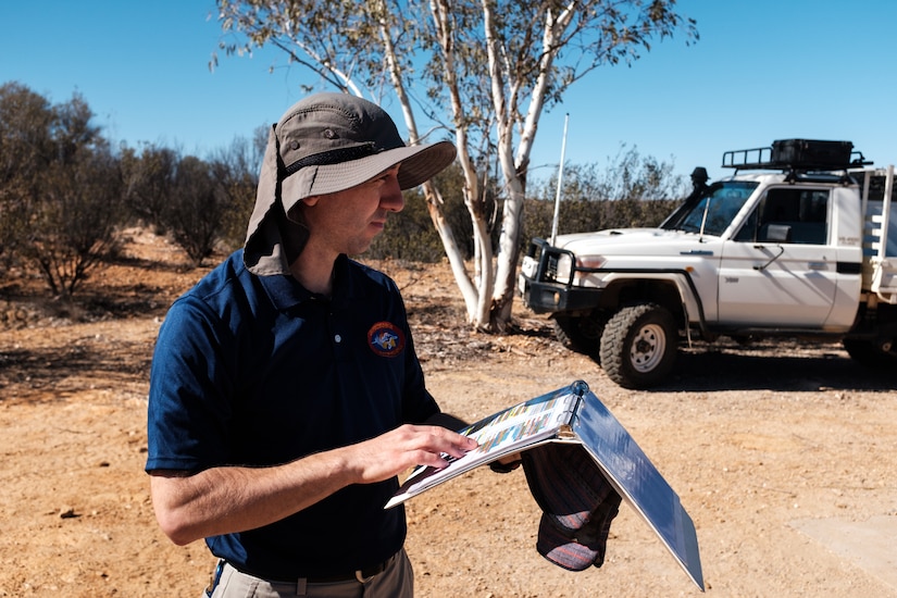 A man holds a binder in the desert near a tree and a truck.