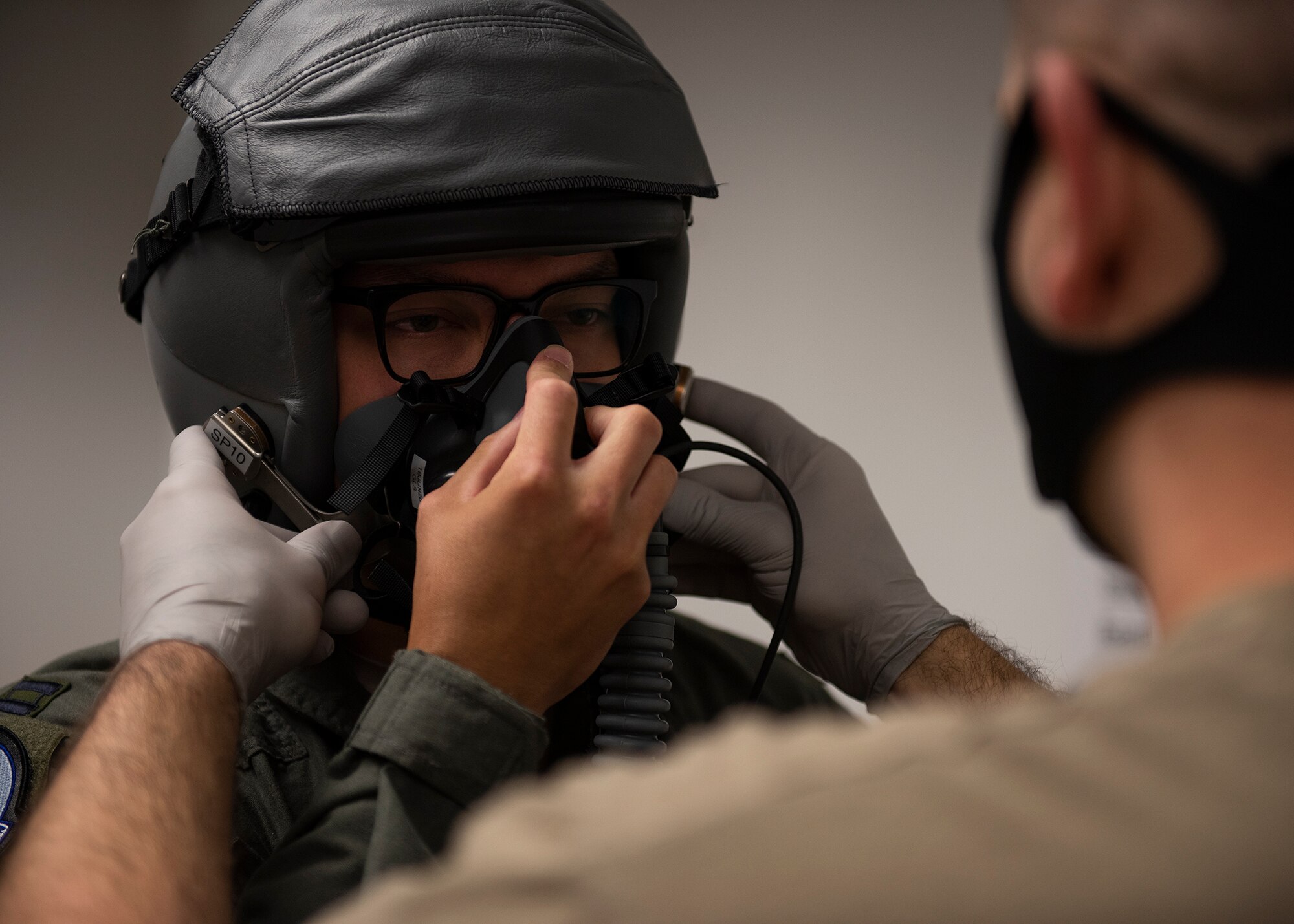 An Airman wearing a helmet holds an oxygen mask to their face while another Airman secures the oxygen mask to the helmet.