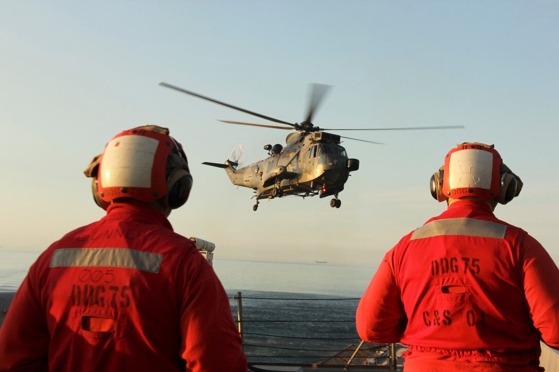 A German navy helicopter flies as two service members watch while standing on a U.S. Navy ship.