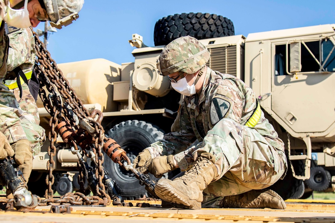 A soldier kneeling on one knee unfastens a metal chain.