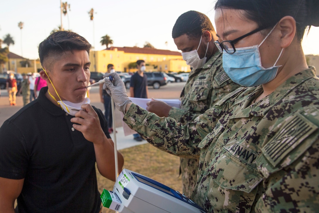 A sailor wearing a mask and gloves holds a thermometer in the mouth of a Marine Corps recruit as others stand nearby outside.