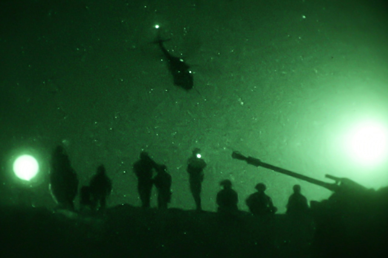 A helicopter flies over troops at night.