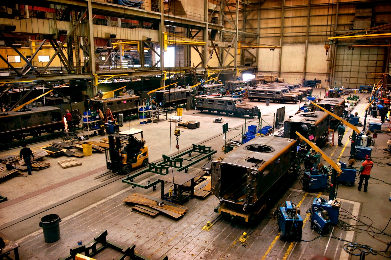 Inside a large manufacturing facility, multiple military vehicles are in various states of assembly.