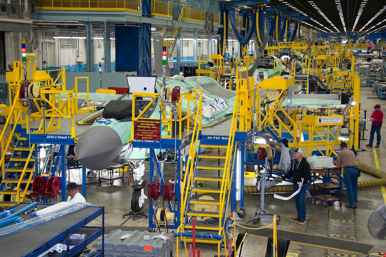 In a large, brightly-lit facility with blue and yellow scaffolding, military aircraft are lined up in varying states of assembly.