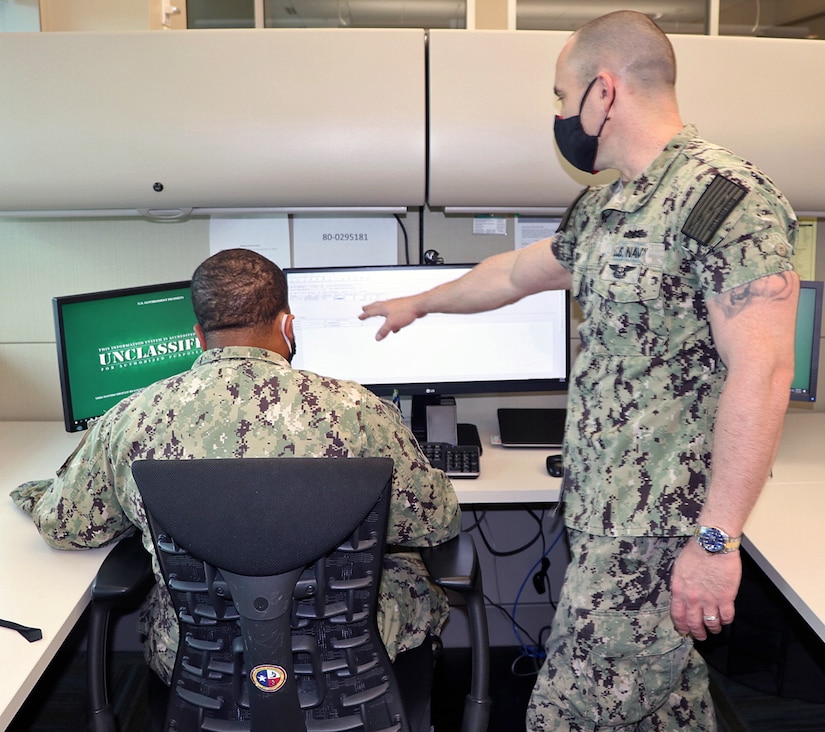 One sailor is seated at a computer monitor as another stands nearby and provides instruction.