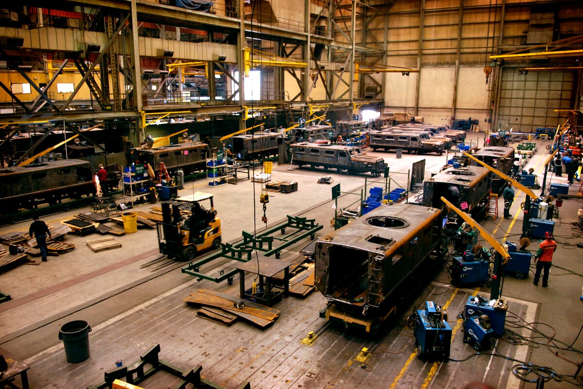 Inside a large manufacturing facility, multiple military vehicles are in various states of assembly.