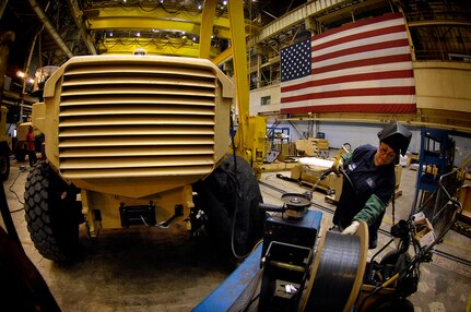 A worker wears a welder’s hood flipped up on her head. She manipulates a roll of wire. A large vehicle sits to her left.