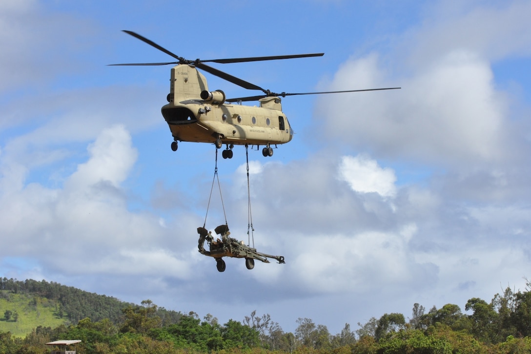 A helicopter carries a howitzer through the air.