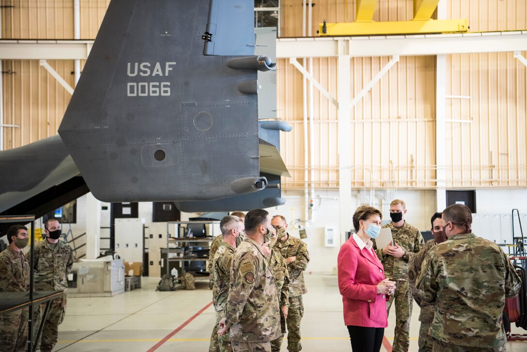 Civilian woman speaks with airmen in a hangar while other airmen work on an aircraft in the background.