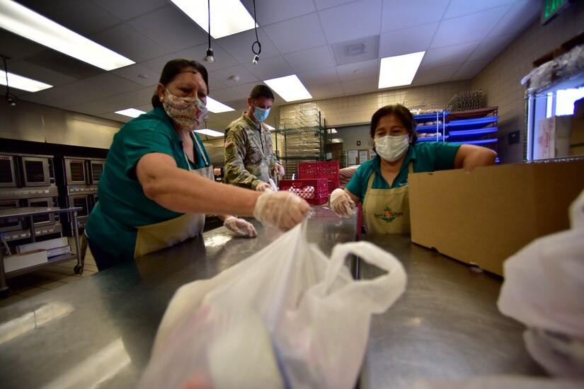 Two women and a soldier, all wearing masks, pack meals for a school lunch program.