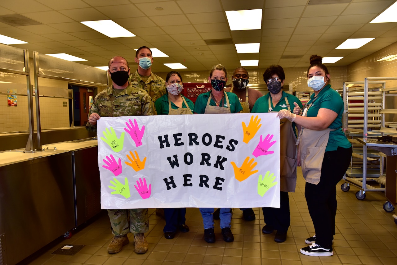 Soldiers and civilians pose for a photo, holding a sign that reads, “Heroes Work Here.”