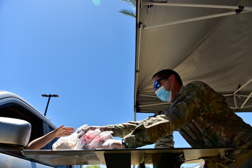 A soldier wearing a face mask uses a tray to give a packaged school lunch to a parent through a car window.