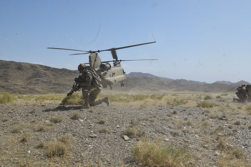 A soldier is on his knee on dusty terrain. Behind him, a CH-47 Chinook lifts off.