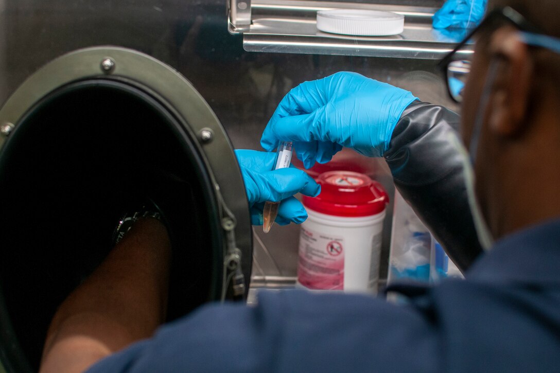 A soldier wearing a face mask and surgical gloves examines a lab specimen.