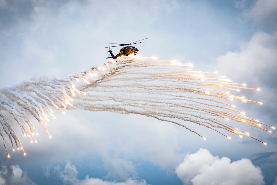 A helicopter flies above a cascade of flares in cloudy blue sky.