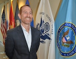 A smiling man stands beside a row of military flags