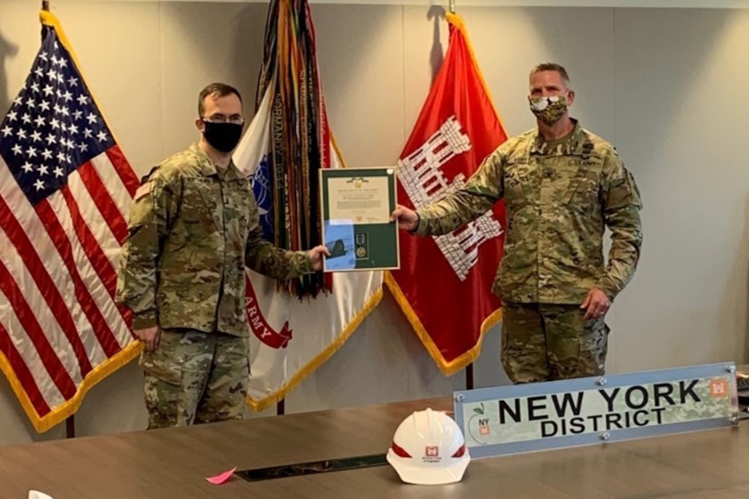 Two soldiers wearing face masks pose in front of an array of flags while holding a military decoration certificate.