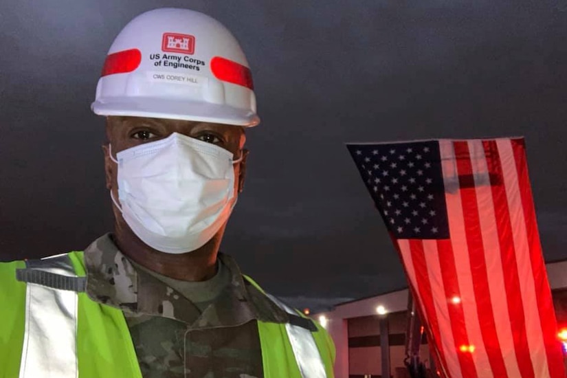 Soldier wearing a white hard hat, yellow reflective vest and a white face mask poses next to a suspended American flag.