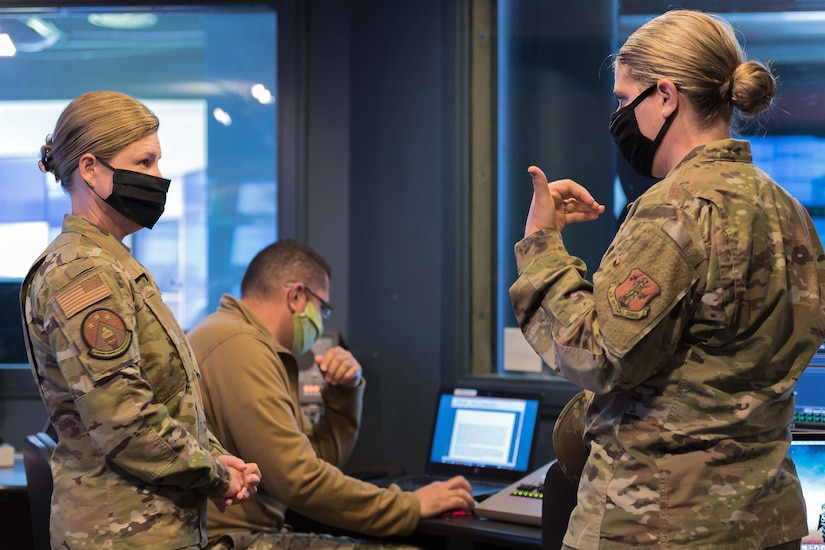 Two airmen wearing face masks talk as a third mask-wearing airman works beside them.