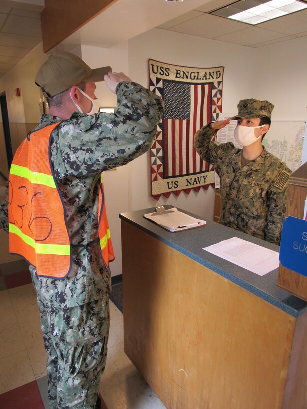 Sailors wearing face masks exchange salutes at a quarterdeck counter.