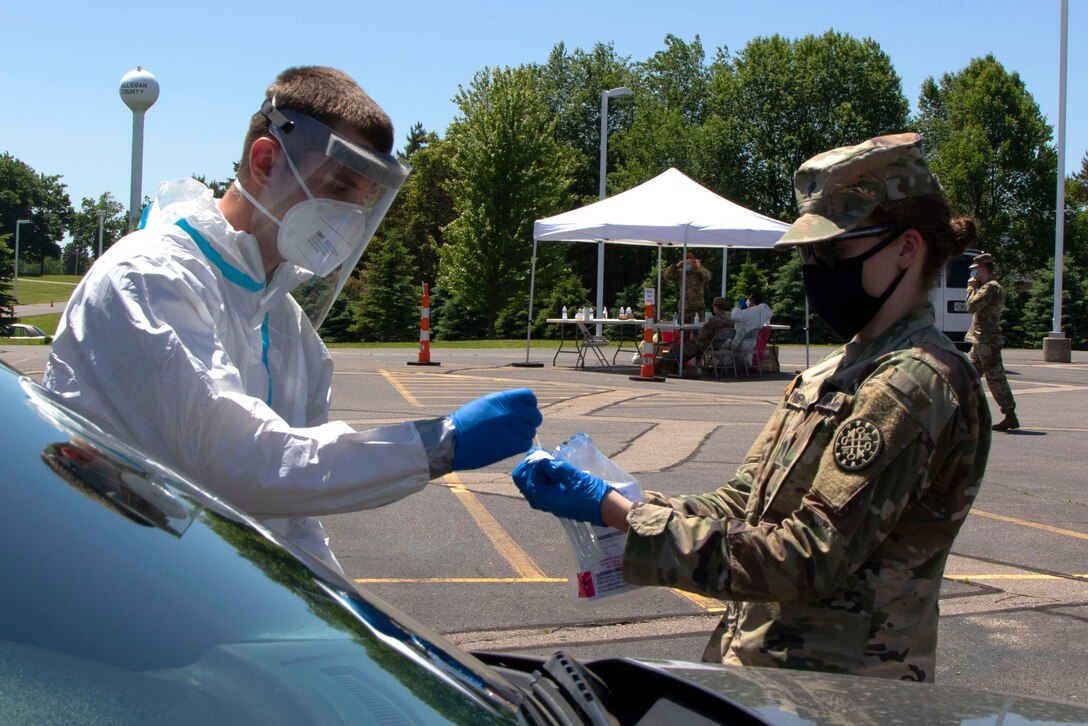 A soldier wearing personal protective equipment puts a COVID-19 test swab in a bag as another soldier holds a bag open