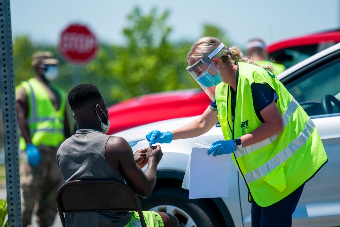 A soldier wearing a medical mask and gloves talks with a person at a drive-thru testing site.