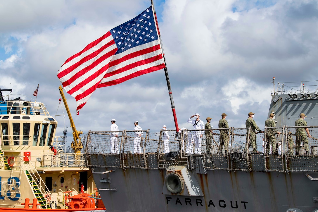 Sailors stand in formation on a ship’s deck as a large American flag waves.
