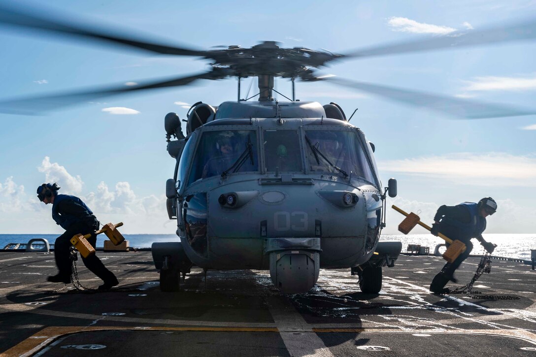 Two sailors remove chocks and chains from a helicopter on the deck of a ship.