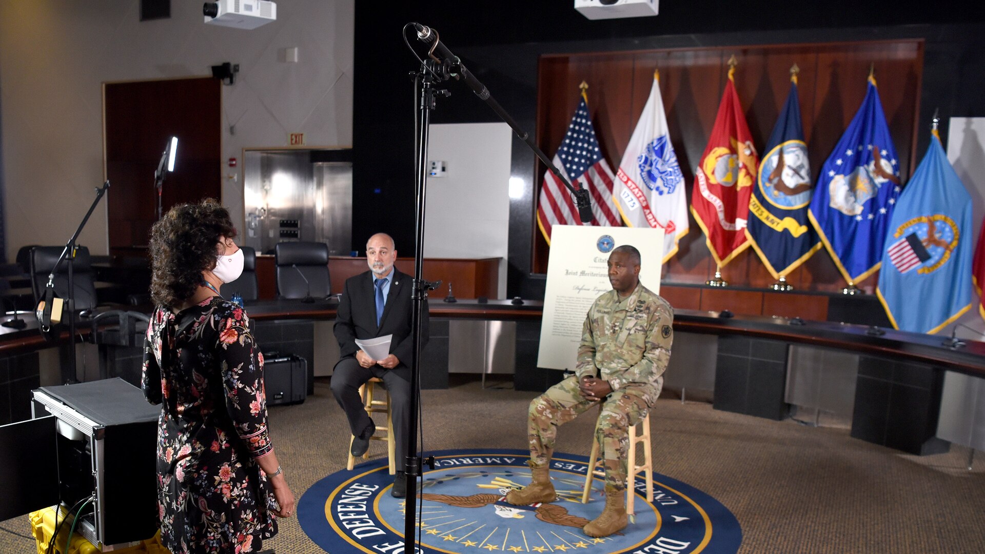 Two men - one white wearing a suit, the other black in an Army uniform - sit on stools with flags behind them as a woman in a white face mask stands in front of them. A tall, arching light illuminates the men.