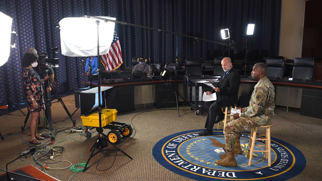 Two men - one white wearing a suit, the other black wearing an Army green camo uniform - sit in stools as a woman wearing a white dress and white face mask stand in front of them. Bright studio lights illuminate the men.