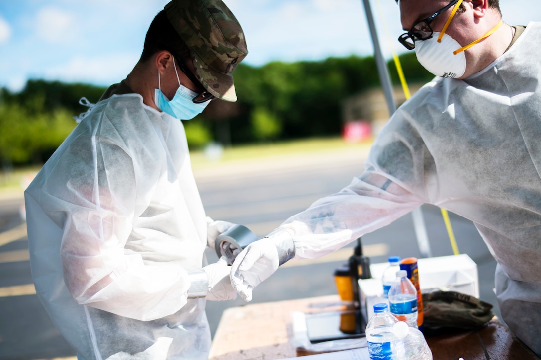 A guardsman wearing personal protective equipment wraps tape around the wrist of another guardsman wearing personal protective equipment.