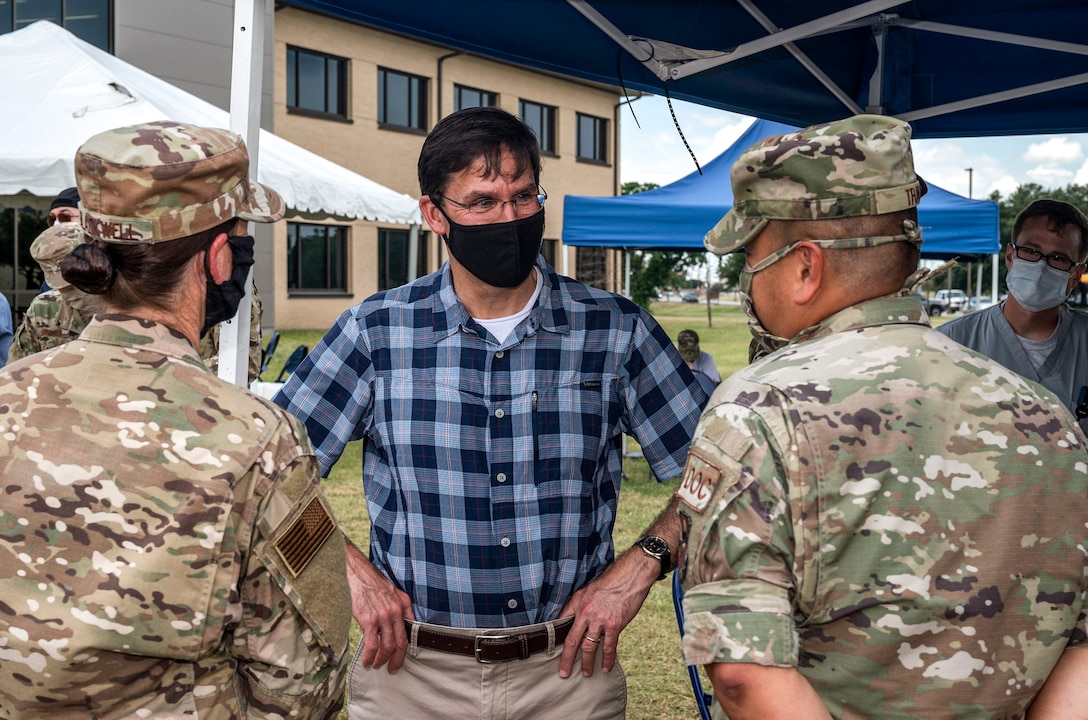 Defense Secretary Dr. Mark T. Esper wears a mask while talking to two airmen outside a building.