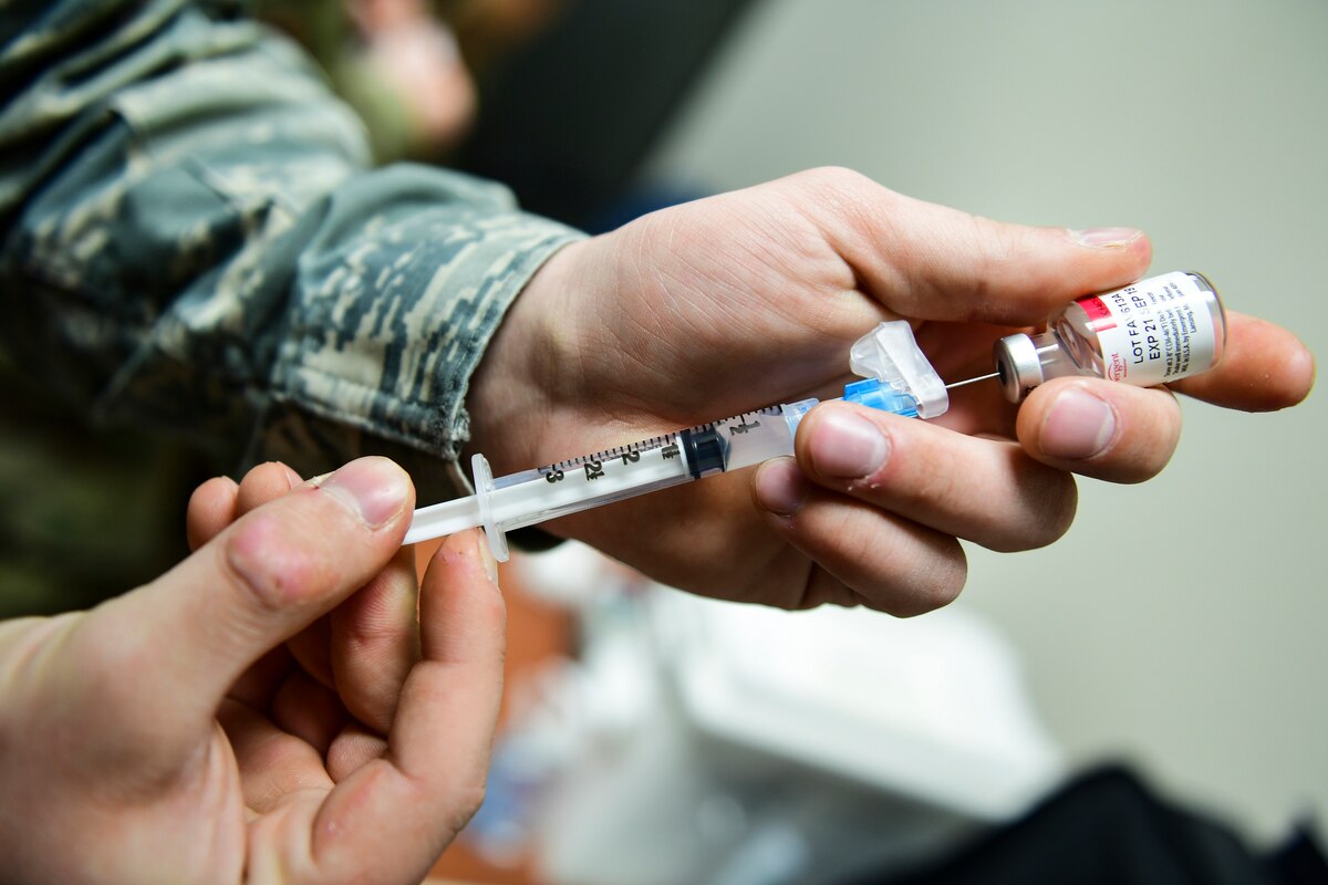 A service member presses a hypodermic needle into a small glass vial.