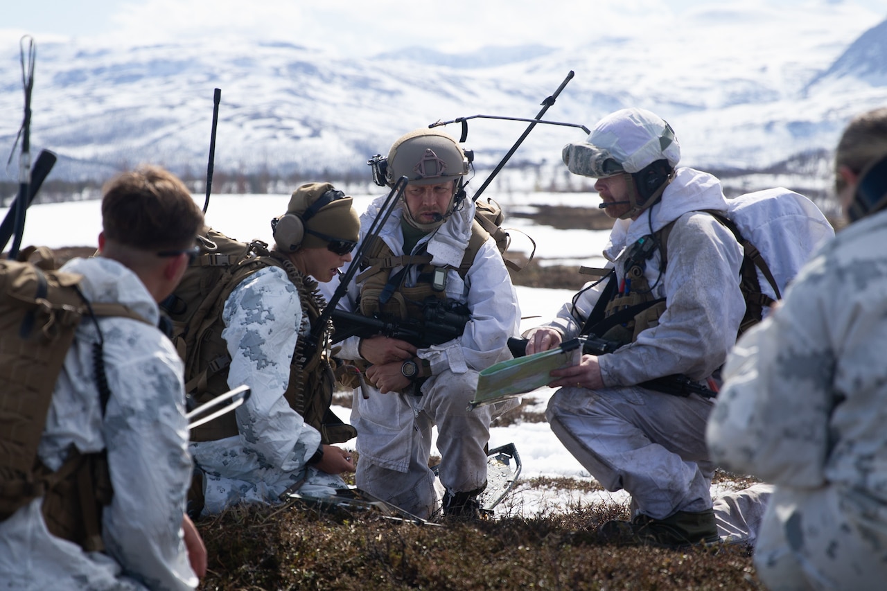 Service members wearing white uniforms kneel in a huddle to examine a map on snowy terrain.