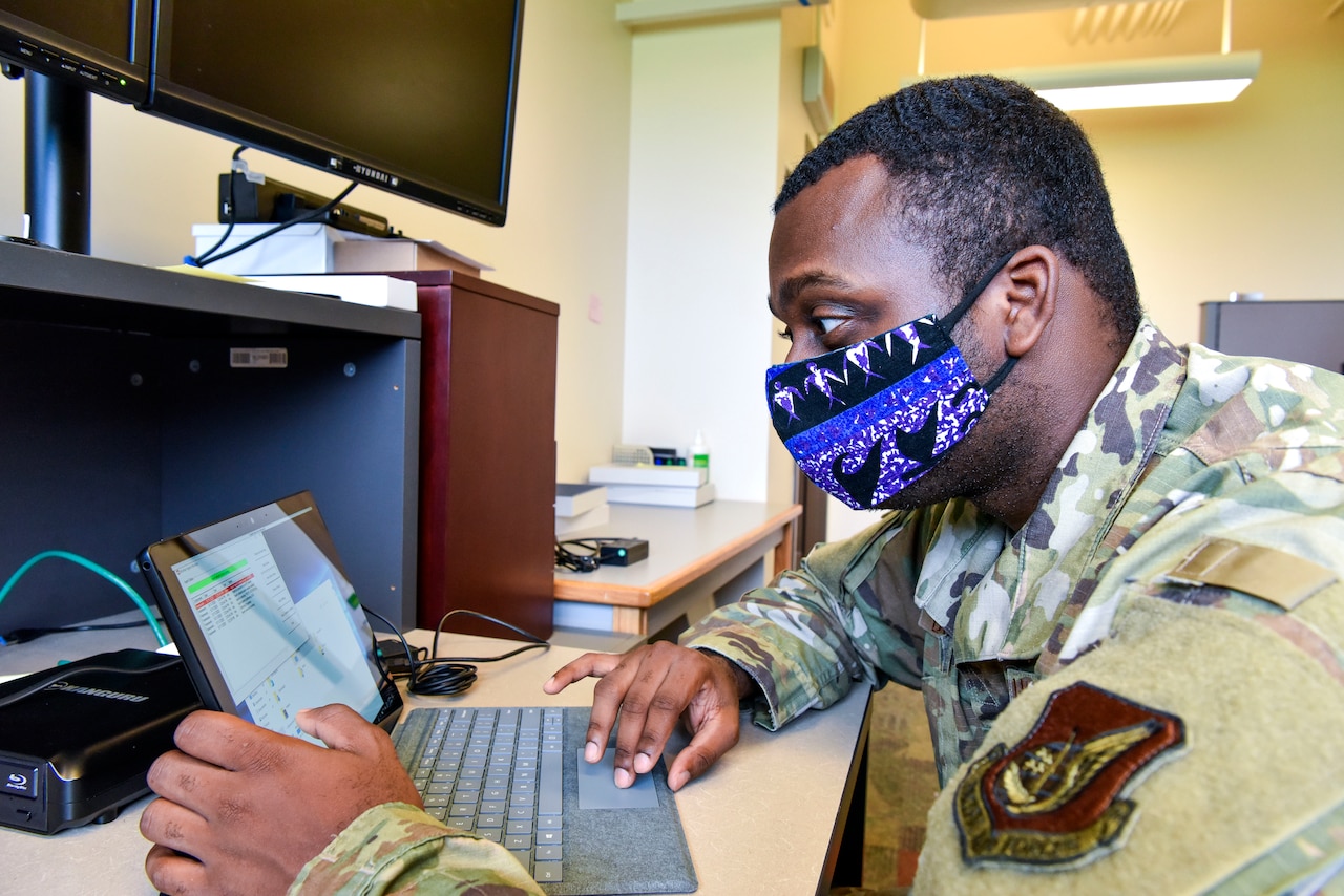 An airman wearing a face mask works on a computer.